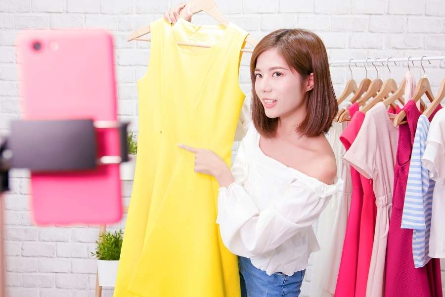 Une jeune femme présente une robe jaune vive devant un smartphone fixé sur un trépied pour vendre des vêtements en ligne.